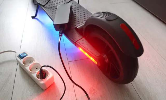 Un chargeur pour trottinette électrique est branché sur une multiprise. La trottinette est éclairée par des LED bleues et rouges.