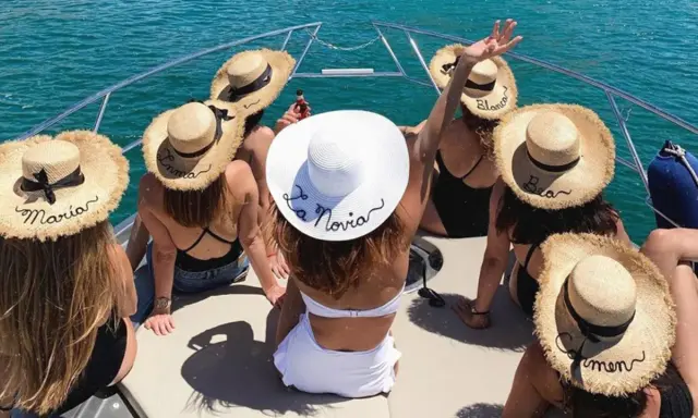 Grupo de amigas en un barco celebrando unas divertidas ideas despedida de soltera. Llevan sombreros de paja con nombres bordados, uno dice "La Novia".