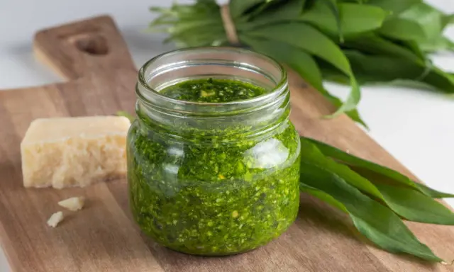 Pesto ail des ours fait maison dans un bocal, avec du fromage et des feuilles fraîches sur une planche en bois.