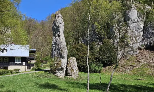 Imponująca skała Igła Deotymy góruje nad zieloną polaną, obok której stoi drewniany domek.
