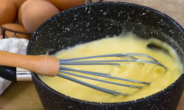 Un fouet mélange une crème pâtissière traditionnelle onctueuse dans une casserole. Des œufs sont visibles en arrière-plan.