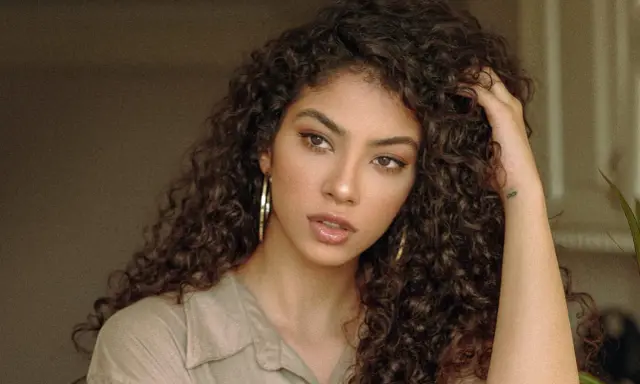 Une jeune femme aux cheveux bruns et bouclés, portant une chemise beige et de grandes boucles d'oreilles. Sa coupe boucles met en valeur son regard intense.