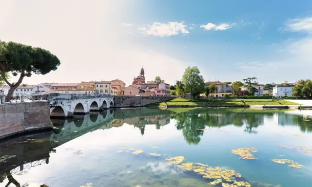 Ponte di Tiberio e il fiume Marecchia a Rimini a settembre, con riflessi sull'acqua e un cielo azzurro con nuvole bianche.