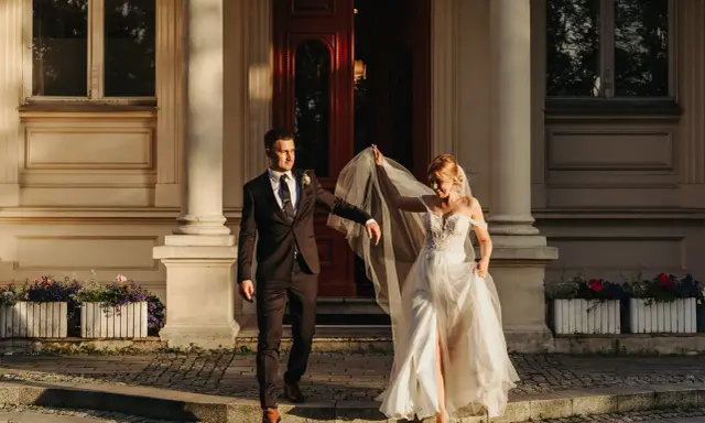 Para młoda podczas sesji zdjęciowej ślubnej schodzi po schodach przed eleganckim budynkiem. Panna młoda w zwiewnej sukni unosi welon, pan młody w garniturze patrzy na nią z uśmiechem.