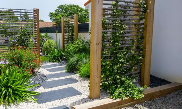 Un chemin de dalles grises serpente à travers un jardin luxuriant. Des treillages en bois servent de support à une plante brise vue grimpante, créant un espace intime.