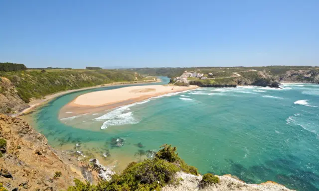 Atemberaubende Aussicht auf Praia de Odeceixe, wo ein Fluss auf den Ozean trifft. Grüne Hügel und ein weißes Dorf säumen die Küste.