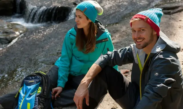 Para, co to jest trekking? Para w czapkach i kurtkach siedzi przy strumieniu, obok plecak.