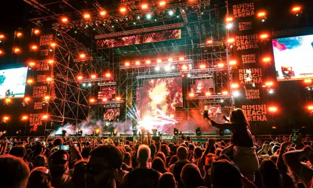 Un concerto elettrizzante alla Rimini Beach Arena. Migliaia di fan in delirio sotto un palco imponente illuminato da luci stroboscopiche e schermi giganti.