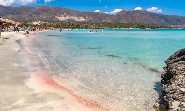 Spiaggia rosa di Creta con bambini che giocano nell'acqua cristallina e montagne sullo sfondo.