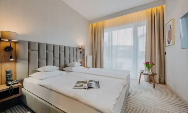 Przytulny executive room z dwoma wygodnymi łóżkami, lampkami nocnymi i dużym oknem z zasłonami. Na stoliku bukiet tulipanów.