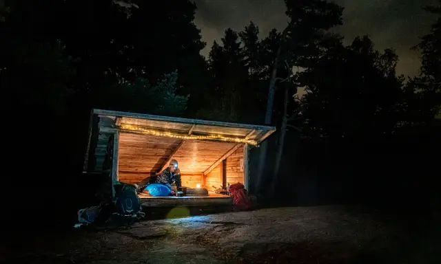 Person myser i ett upplyst vindskydd i skogen på natten. Ryggsäckar står utanför.