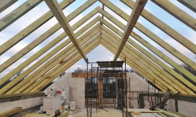 Konstrukcja dachu z drewnianych belek, widoczna ściana szczytowa z bloczków, rusztowania i materiały budowlane.