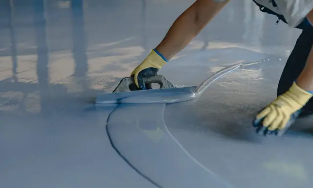 Persona con guantes amarillos aplica una capa de pintura azul clara sobre un suelo de terrazo con una llana.