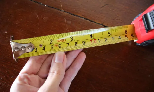 Una mano sujeta una cinta métrica amarilla sobre una mesa de madera. La cinta muestra medidas en pies y metros, demostrando para que sirve la cinta métrica: medir distancias.