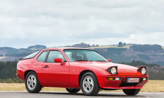 Roter Porsche 925 mit ausklappbaren Scheinwerfern steht auf einer Straße, im Hintergrund bewaldete Hügel.