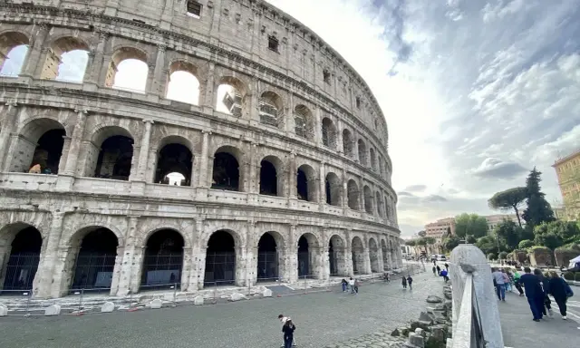 Le Colisée, monument emblématique de Rome, se dresse majestueusement sous un ciel nuageux. Des visiteurs explorent ses environs pavés.