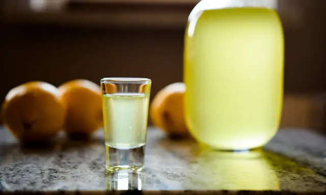 Szklaneczka domowego limoncello, obok cytryny i dużej butelki. Idealny przepis na orzeźwiający trunek.