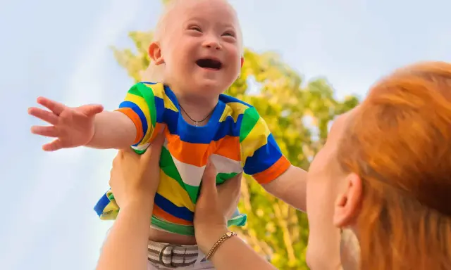 Niño con síndrome de Down, sonriente, es levantado por una persona. Sus rasgos son visibles y llenos de alegría.