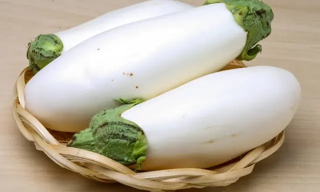 Trois aubergines blanches, d'une blancheur immaculée, reposent dans un panier en osier tressé sur un fond en bois clair.