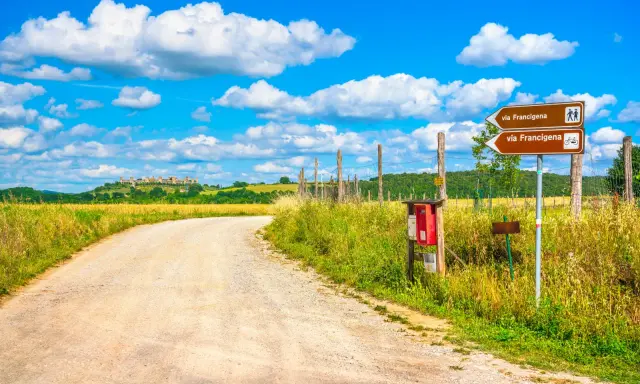 Segnaletica per la Via Francigena, un antico cammino. La strada sterrata conduce verso un borgo collinare sotto un cielo azzurro con nuvole bianche.