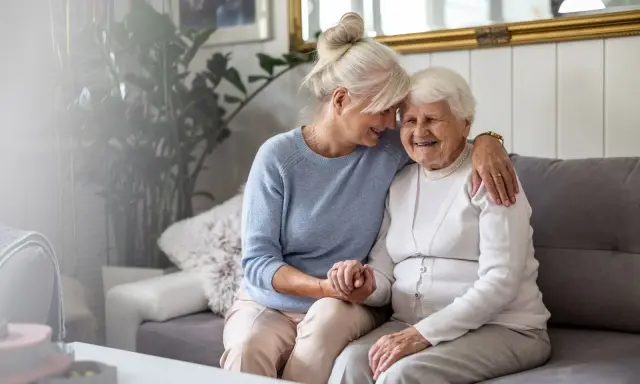 Deux femmes âgées s'enlacent tendrement sur un canapé. L'une d'elles semble visiter sa mère, une belle dame, dans sa maison en EHPAD.