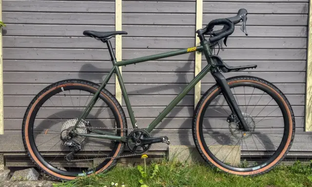 Grünes Rose Hobo Test Gravelbike mit braunen Reifen und Gepäckträger vor Holzwand.