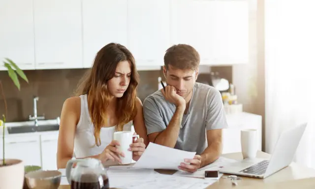 Couple pensif devant des papiers, peut-être une attestation d'assurance villégiature, dans une cuisine lumineuse.