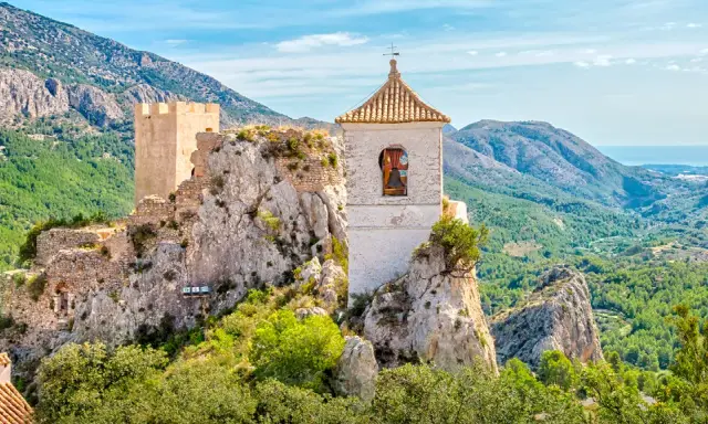 Das Castell de Guadalest thront auf einem Felsen, umgeben von grünen Bergen und dem blauen Meer in der Ferne. Ein Glockenturm ragt empor.