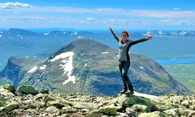 Kvinna med armarna utsträckta på en stenig topp, njuter av utsikten över fjällandskap med snöfläckar. Ett perfekt berg att bestiga!