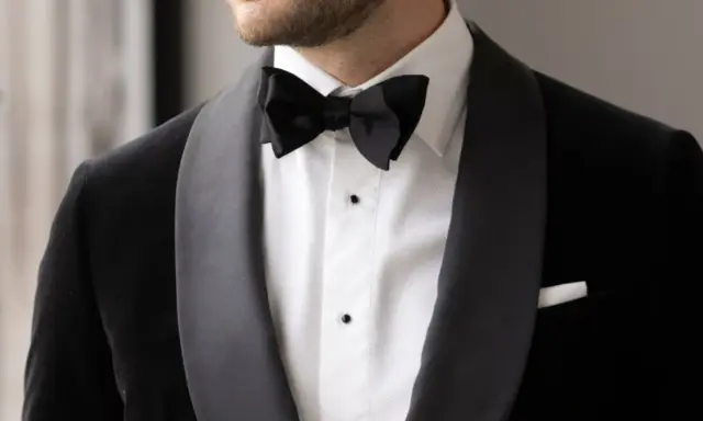 A man wears a sharp black suit with a crisp white shirt and a black bow tie, ready for a formal occasion.