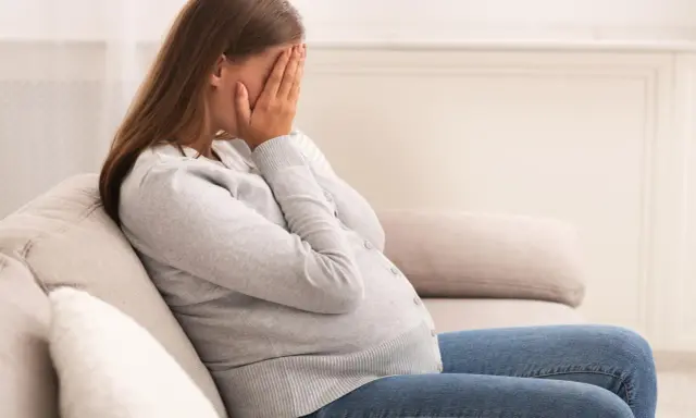 Kobieta w ciąży siedzi na kanapie, zakrywając twarz dłońmi. Jej postawa wyraża głęboki smutek, być może związany z diagnozą wady letalnej.
