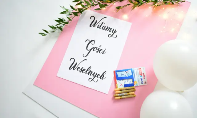 Tablica powitalna wesele z napisem "Witamy Gości Weselnych", ozdobiona gałązką zieleni i lampkami. Obok taśma, złote pisaki i białe balony.