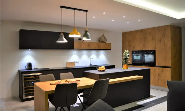 Une cuisine moderne aux accents beige et noir, avec un îlot central, des meubles en bois et des suspensions colorées.