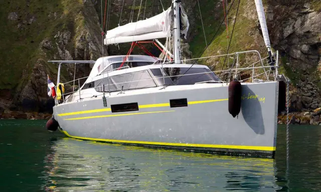 Le voilier "Bi-loup 109" gris avec des bandes jaunes, ancré près d'une falaise verdoyante.