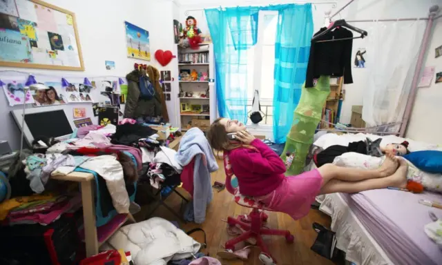 Chica en su habitación desordenada, hablando por teléfono, rodeada de ropa y objetos esparcidos.