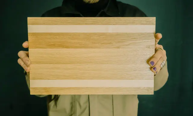 Hände halten ein rechteckiges Schneidebrett aus naturbelassenen holzbrettern mit hellen und dunkleren Streifen.