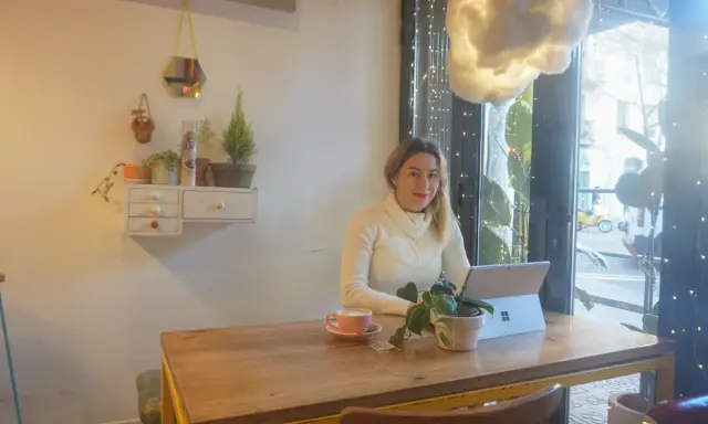 Kobieta pracuje na laptopie w kawiarni, ciesząc się kawą i roślinami. To idealne miejsce na dodatkową pracę zdalną.