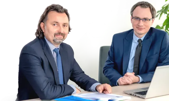 Dwóch mężczyzn w garniturach siedzi przy biurku z dokumentami i laptopem.