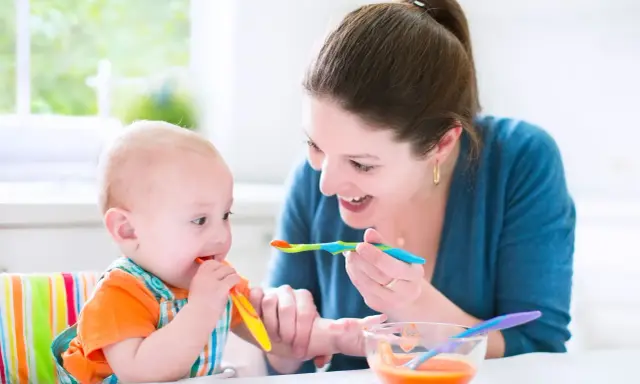 Uśmiechnięta mama karmi łyżeczką 7 miesięczne dziecko, które samo próbuje jeść.