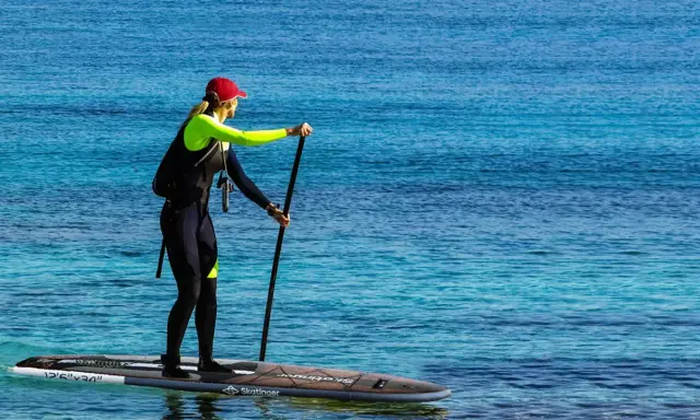 Kobieta w piance i czapce na desce sup, z wiosłem w ręku. Widać leash sup.