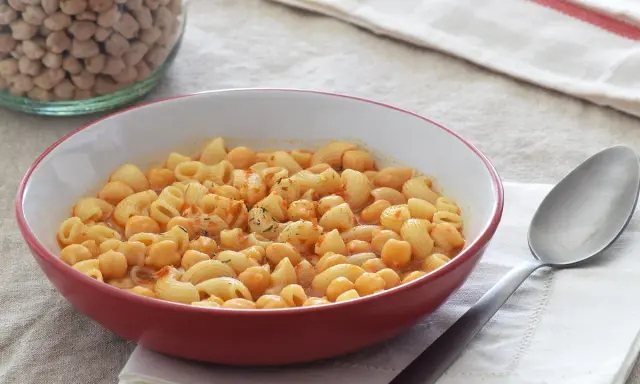 Un plato humeante de pasta e ceci, con garbanzos y macarrones en una salsa rojiza, junto a un tarro de garbanzos secos.