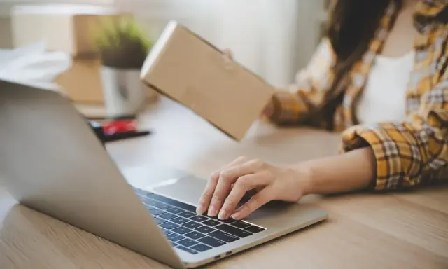 Osoba pakuje paczkę, sprawdzając na laptopie, kto ponosi koszty zwrotu towaru.