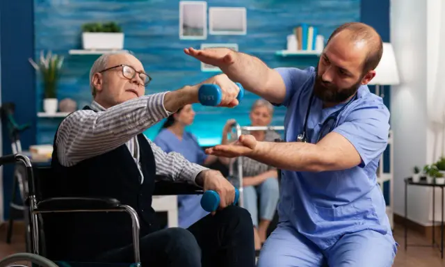 Starszy mężczyzna w wózku wykonuje ćwiczenia z hantlem pod okiem fizjoterapeuty. To kluczowy element rehabilitacji ręki po udarze.