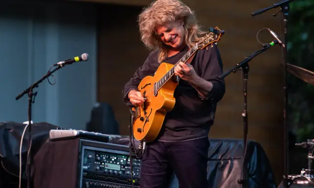 Muzyk z burzą blond włosów gra na gitarze, tworząc wspaniały śpiew w duecie z niewidocznym wokalistą.