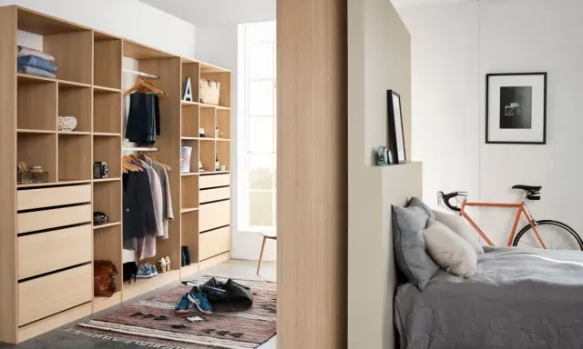 Un armario moderno y funcional, ideal para aprender como organizar un armario. Ropa colgada, cajones y estantes ordenados, con una bicicleta naranja y una cama al fondo.
