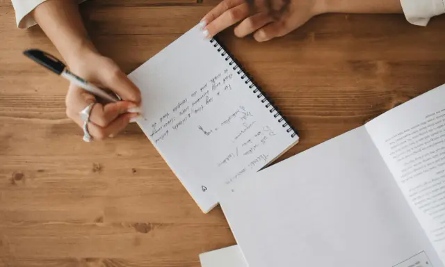 A person's hands are writing notes in a notebook. The notes mention "Theme: coronavirus", "self-isolation", and "lockdown".