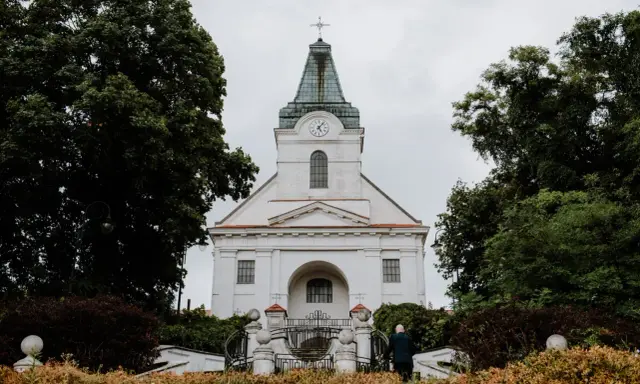 Biały kościół z zegarem na wieży, otoczony drzewami. Zastanawiasz się, jak sprawdzić do której parafii należę?