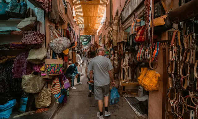 Bunte Taschen und Gürtel hängen in einem engen Gang des Marrakesch Basars. Ein Mann geht mit einer blauen Tüte durch das bunte Treiben.