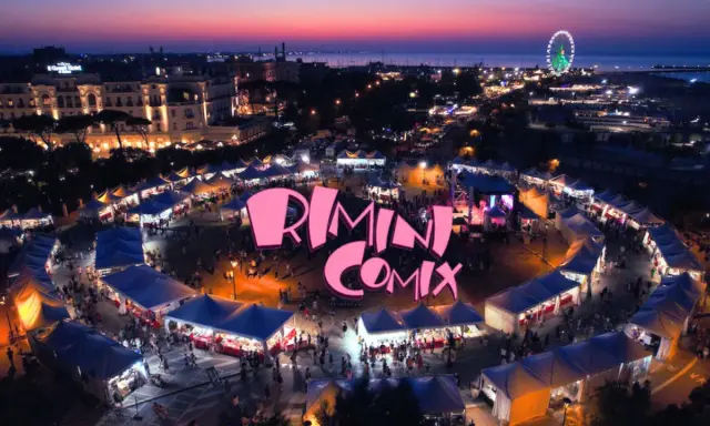 Un vivace evento serale a Rimini Comix, con bancarelle illuminate, una ruota panoramica e il logo rosa che domina la scena.
