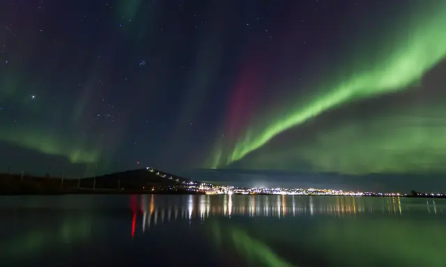 Gröna och lila norrsken dansar över en stjärnklar himmel ovanför Kiruna. Stadens ljus speglas i det lugna vattnet.